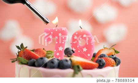 Birthday cake number 79, pink candle on beautiful cake with berries and lighter with fire against background of white clouds and pink sky. Close-up 78089314