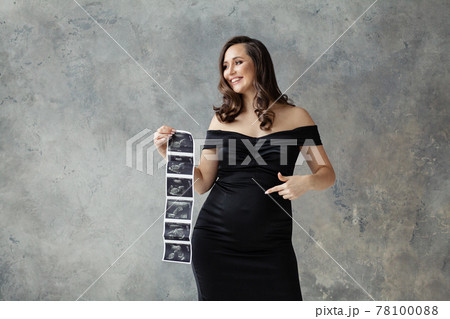 Pregnant woman pointing at her tummy and showing ultrasound scan image on gray 78100088
