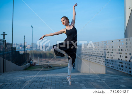 Ballerina in a tutu posing next to the fence. Beautiful young woman in black dress and pointe dancing over city background. Portrait of a gorgeous ballerina performing a dance outdoors 78100427