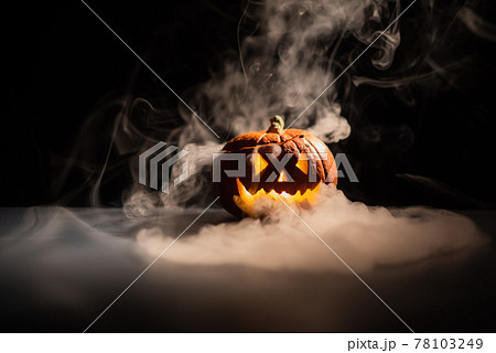 Halloween, orange pumpkin with a scary luminous face on a dark background. Thick gray smoke comes out and spreads across the black table. A close-up of a flashlight on the eve of all the saints Halloween, orange pumpkin with a scary luminous face on a dark background. Thick gray smoke comes out and spreads across the black table. A close-up of a flashlight on the eve of all the saints 78103249