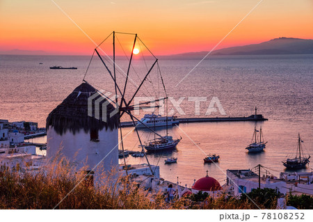 Famous traditional Greek windmill over port of Mykonos, Greece at sunset. Beautiful sky, sun touch sea horizon, boats, pier, high grass in foreground. 78108252