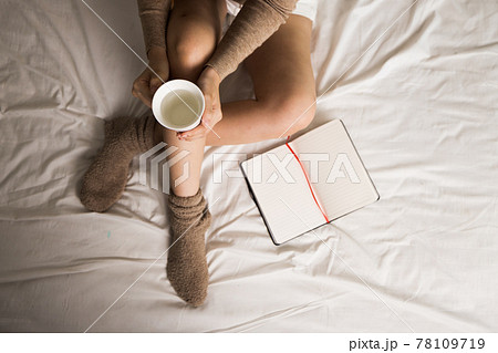 Top view of woman sitting on her bed with her notebook open beside her. She is also holding a cup of water in her hand. She wear wool sock. Top view of woman sitting on her bed with her notebook open beside her. She is also holding a cup of water in her hand. She wear wool sock. 78109719