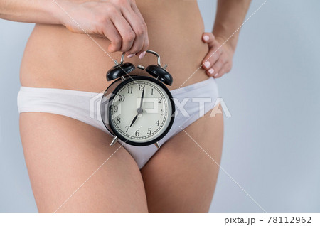 A faceless woman in shorts holds an alarm clock on a white background. Female biological clock concept 78112962