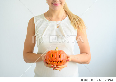 A woman in a white blouse holds a pumpkin for Halloween. Close-up of female hands with a lantern jack on a white background on the eve of All Saints. foreground. Trick or treat. Scary grimace. 78112998