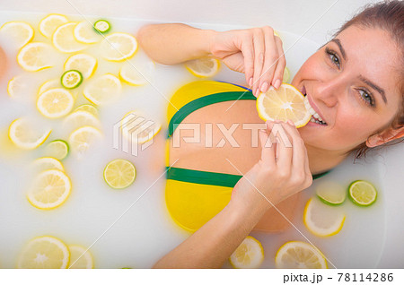 Beautiful young red-haired woman takes a bath with milk and citrus fruits. Portrait of Caucasian smiling girl eating lemon wedge in skin whitening bath. Taking care of the body. 78114286
