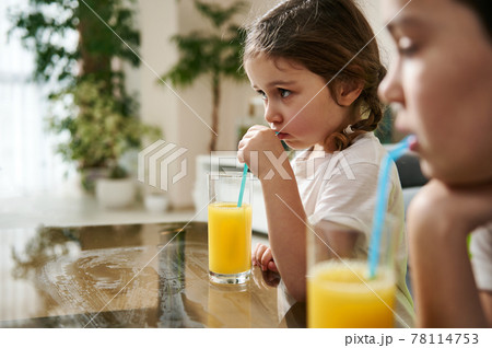 Beautiful baby girl drinking orange juice. Blurry boy on the foreground 78114753