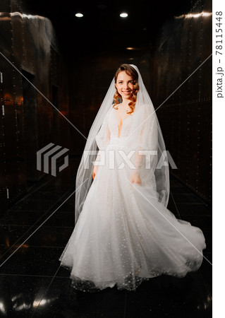 beautiful bride in a white elegant dress, with a veil goes down a dark corridor beautiful bride in a white elegant dress, with a veil goes down a dark corridor 78115448