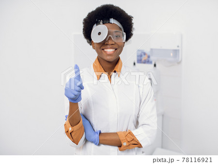 Portrait of young afro american lady otolaryngologist with a medical mirror on her head, smiling and looking at the camera, showing her thumb up. Female ENT doctor shows thumb up at modern clinic. Portrait of young afro american lady otolaryngologist with a medical mirror on her head, smiling and looking at the camera, showing her thumb up. Female ENT doctor shows thumb up at modern clinic. 78116921