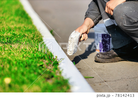 A worker in overalls paints a border with white paint on a summer day. Urban services, landscaping. 78142413