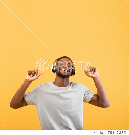 Excited black guy listening to music in headphones and raising fingers up at free space, yellow background, crop Excited black guy listening to music in headphones and raising fingers up at free space, yellow background, crop 78142886