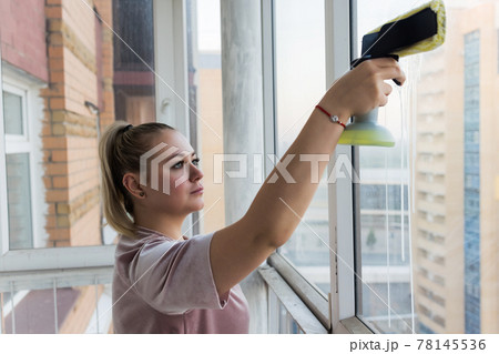 Beautiful young housewife in working clothes washing windows by professional tools at home. 78145536