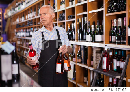 Confident elderly salesman of wine house arranging wine bottles on shelves rack Confident elderly salesman of wine house arranging wine bottles on shelves rack 78147165