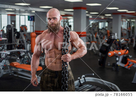 A bald, bearded bodybuilder with large relief muscles holds a heavy thick chain in his hands. Bright gym. 78166055
