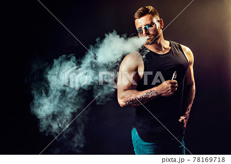 Bearded brutal male in sunglasses smoking a vapor cigarette as an alternative to tobacco. Studio shot on dark background. 78169718