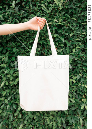 Blank white tote bag canvas fabric with handle mock up design. Close up of woman hand holding eco or reusable shopping bag on green leaves background. No plastic bag and ecology concept. Copy space. 78173881