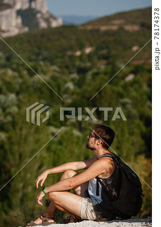 Young male hiker with backpack relaxing on top of a mountain during calm summer sunset 78174378