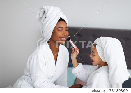 Beautiful black mother and daughter in bath robes and towels applying cosmetics on bed at home Beautiful black mother and daughter in bath robes and towels applying cosmetics on bed at home 78180360