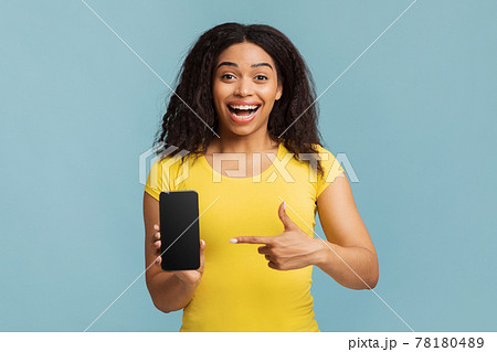 Joyful black lady pointing at smartphone with blank screen on blue studio background, mockup for mobile app design 78180489