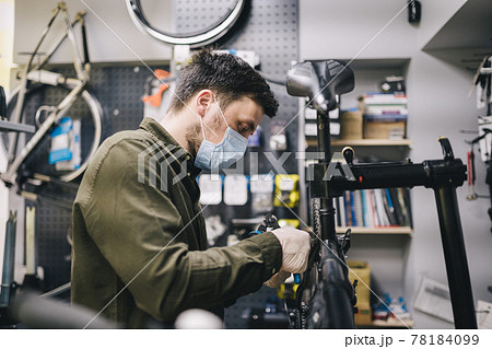 Bicycle mechanic wearing protective mask and gloves repairs customers bicycle wheel in accordance with quarantine standards during coronavirus pandemic. Small business during covid 19 lockdown 78184099