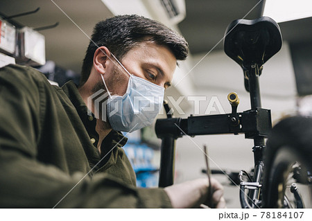 Bicycle shop repairman works in bicycle service and repair workshop during coronavirus quarantine wearing face shield and gloves, new norm. Mechanic repairing bicycle wheel wearing medical mask 78184107