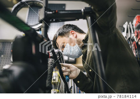 Bicycle mechanic wearing protective mask and gloves repairs customers bicycle wheel in accordance with quarantine standards during coronavirus pandemic. Small business during covid 19 lockdown 78184114