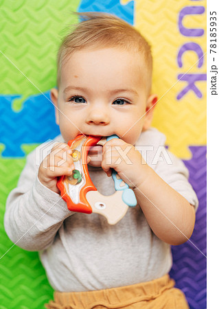 Tiny baby in a white bodysuit holds and gnaws a bright rattle lying on a colored rug Tiny baby in a white bodysuit holds and gnaws a bright rattle lying on a colored rug 78185935