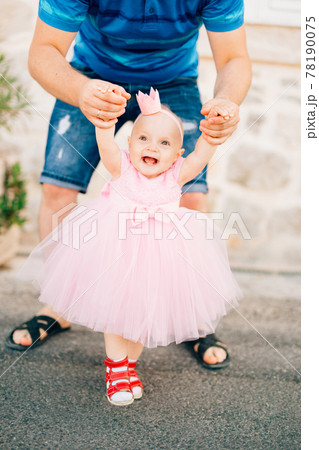 A charming little girl in a pink dress, sneakers and with a crown on her head holding dad by the hands and laughing A charming little girl in a pink dress, sneakers and with a crown on her head holding dad by the hands and laughing 78190075
