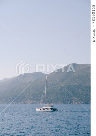 White yacht catamaran with lowered sails sails on the sea against the background of mountains. White yacht catamaran with lowered sails sails on the sea against the background of mountains. 78190158