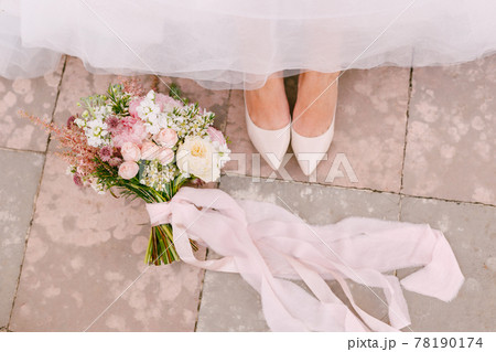 The bride's legs in graceful white shoes peek out from under the skirt, next to the bride's bouquet lies, close-up The bride's legs in graceful white shoes peek out from under the skirt, next to the bride's bouquet lies, close-up 78190174