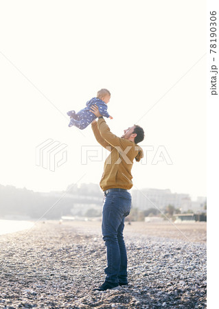 Smiling dad in a brown jacket and jeans throws a small child in a blue overalls high on his arms while standing on a pebble beach 78190306