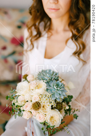 A bride in a lace robe sits on a sofa in a hotel room and holds a wedding bouquet in her hands, close-up A bride in a lace robe sits on a sofa in a hotel room and holds a wedding bouquet in her hands, close-up 78190420