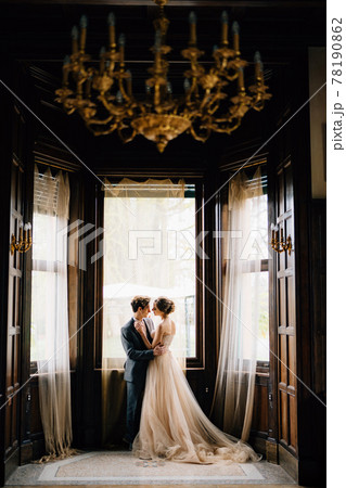 Groom almost kisses bride against the background of the large windows of the old villa. View from the next room with a chandelier with candlesticks on the ceiling. Lake Como Groom almost kisses bride against the background of the large windows of the old villa. View from the next room with a chandelier with candlesticks on the ceiling. Lake Como 78190862