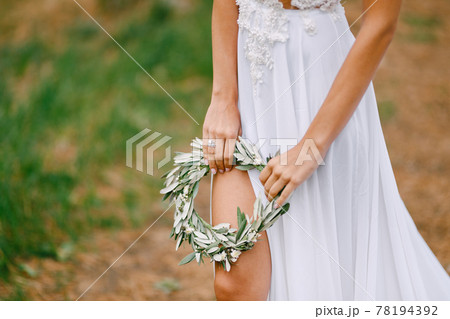 Beautiful bride in a white embroidered dress holds a wreath of olive leaves. Bride's leg is visible through the cut of the dress 78194392
