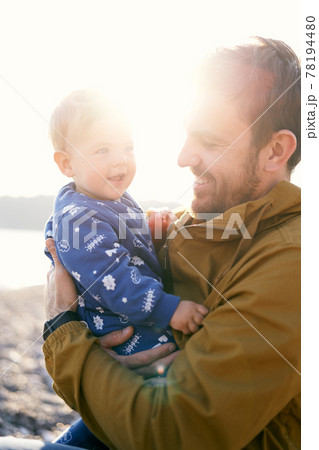 Smiling dad in a brown jacket sits and holds in his arms a small smiling child in a blue overalls. Sun shines brightly. Close-up Smiling dad in a brown jacket sits and holds in his arms a small smiling child in a blue overalls. Sun shines brightly. Close-up 78194480