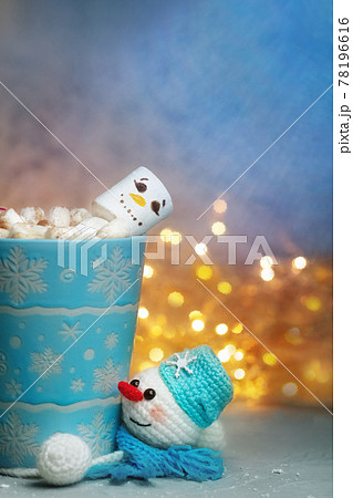 winter mug with marshmellow snowman and knitted handmade stand snowman in scarf against background of festive lights winter mug with marshmellow snowman and knitted handmade stand snowman in scarf against background of festive lights 78196616