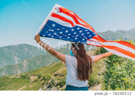 Girl in medical mask holding an American flag in her hands. Celebrate the fourth of July during pandemic Girl in medical mask holding an American flag in her hands. Celebrate the fourth of July during pandemic 78200512