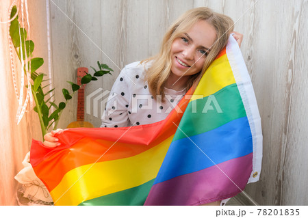 Young blonde millennial hippie woman with rainbow LGBTQ flag at home balcony. Peace and freedom the symbol of LGBT, spending and sharing loving time. Support LGBTQ community. 78201835