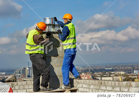 Two workers in protective clothing and safety helmets are installing a pipe for ventilation system on roof of modern building under construction Two workers in protective clothing and safety helmets are installing a pipe for ventilation system on roof of modern building under construction 78206927