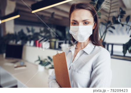 Young businesswoman in face mask standing in office and holding clipboard with documents 78209423