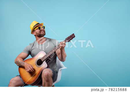 Handsome bearded guitarist in yellow hat and sunglasses sitting on chair and singing while playing on acoustic guitar isolated on blue background Handsome bearded guitarist in yellow hat and sunglasses sitting on chair and singing while playing on acoustic guitar isolated on blue background 78215688