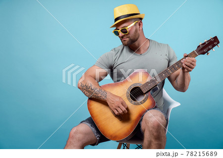 Handsome bearded guitarist in yellow hat and sunglasses sitting on chair and singing while playing on acoustic guitar isolated on blue background 78215689