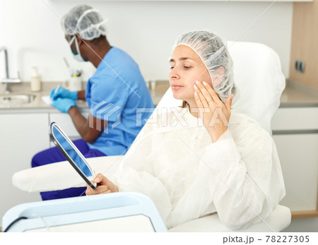 Young woman sitting in armchair and looking to mirror before mesotherapy 78227305
