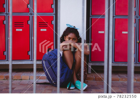 Unhappy african american schoolgirl sitting by lockers in school corridor with schoolbag Unhappy african american schoolgirl sitting by lockers in school corridor with schoolbag 78232550