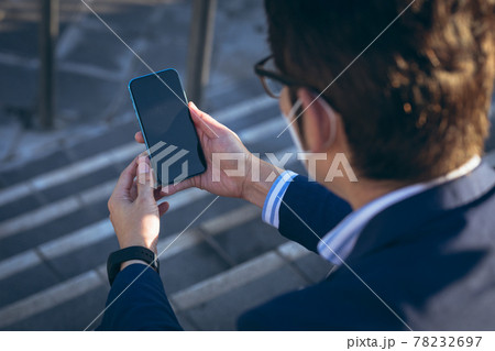 Asian businessman wearing face mask using smartphone sitting on steps in city street Asian businessman wearing face mask using smartphone sitting on steps in city street 78232697