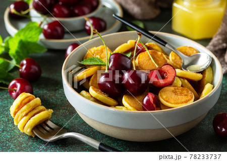 Trendy home breakfast with tiny pancakes cereal close-up. Mini pancakes, honey and cherries on a stone table. Trendy home breakfast with tiny pancakes cereal close-up. Mini pancakes, honey and cherries on a stone table. 78233737