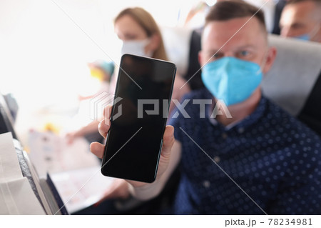 Man in medical protective mask in aircraft cabin holds smartphone with screen forward Man in medical protective mask in aircraft cabin holds smartphone with screen forward 78234981