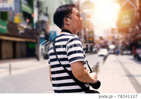 Side rear view of handsome young male tourist with spectacles walking and roaming around the city with camera and side bag while looking away Side rear view of handsome young male tourist with spectacles walking and roaming around the city with camera and side bag while looking away 78241137