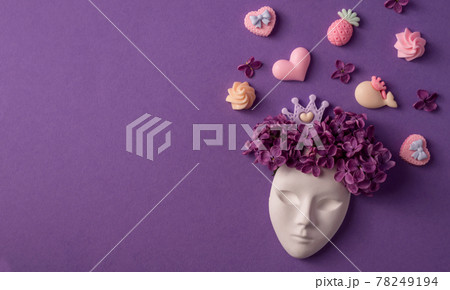 Plaster face decorated with lilac flowers, crown and sweets. Lilac as hairstyle on the head figurine against purple background. Flat lay composition. 78249194