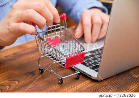 A man sits in a shirt at a laptop and holds a trolley. The concept of wholesale purchases on the Internet. A man sits in a shirt at a laptop and holds a trolley. The concept of wholesale purchases on the Internet. 78268026