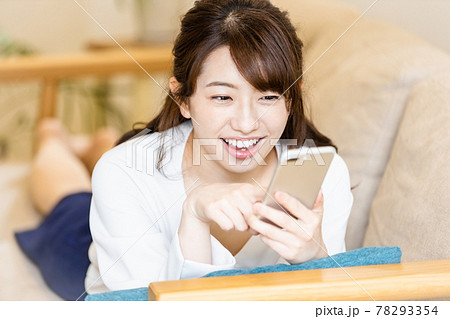 リビングのソファに寝そべってスマホの画面を見る女性 78293354